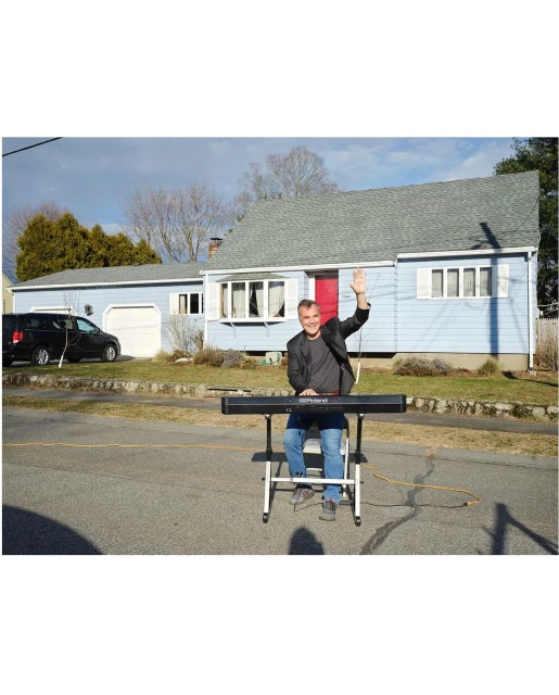 Adult male playing keyboard outside in residential street, gesturing with his left hand, with a house and parked car visible behind him.