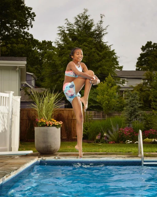 Young girl in swimwear jumps over an outdoor swimming pool, with lush green trees and plants visible in the background on a bright day.