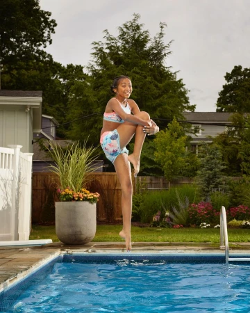 Young girl in swimwear jumps over an outdoor swimming pool, with lush green trees and plants visible in the background on a bright day.