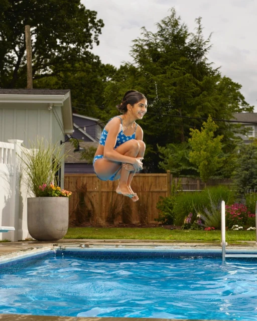 A young girl in a blue swimsuit jumps over a blue outdoor swimming pool with green foliage, a white fence, and a house visible.