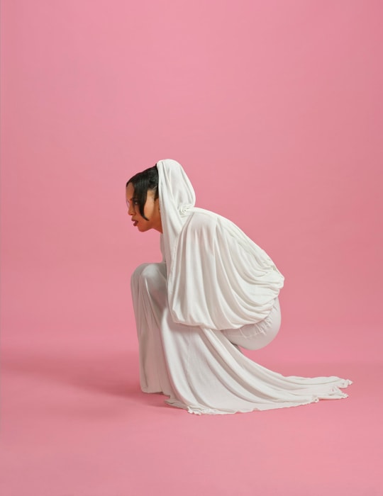 An Asian female model in a white hooded dress crouches on a pink seamless backdrop, showcasing a fashion-forward and minimalist style.