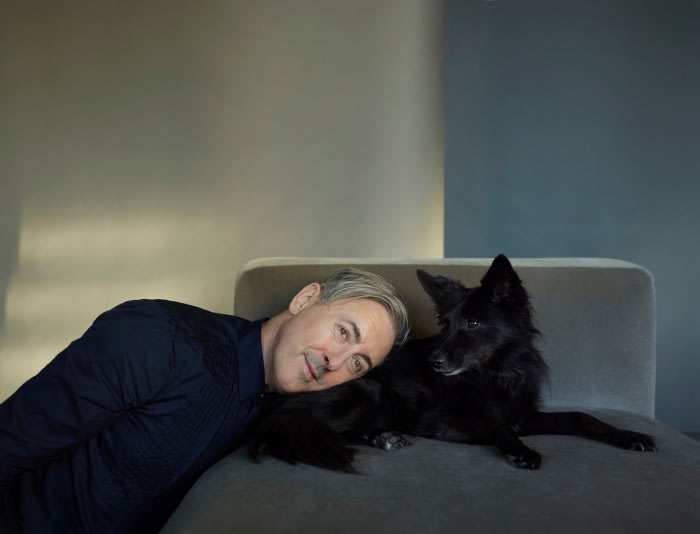 A man rests his head near a long-haired black dog on a gray chaise lounge indoors against a wall with gray and blue sections.