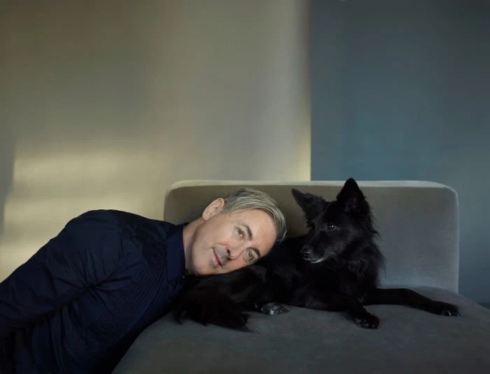 A man rests his head near a long-haired black dog on a gray chaise lounge indoors against a wall with gray and blue sections.
