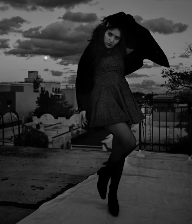 Monochrome portrait of a woman posing on a rooftop against a cloudy sky, wearing a dark coat and dress.