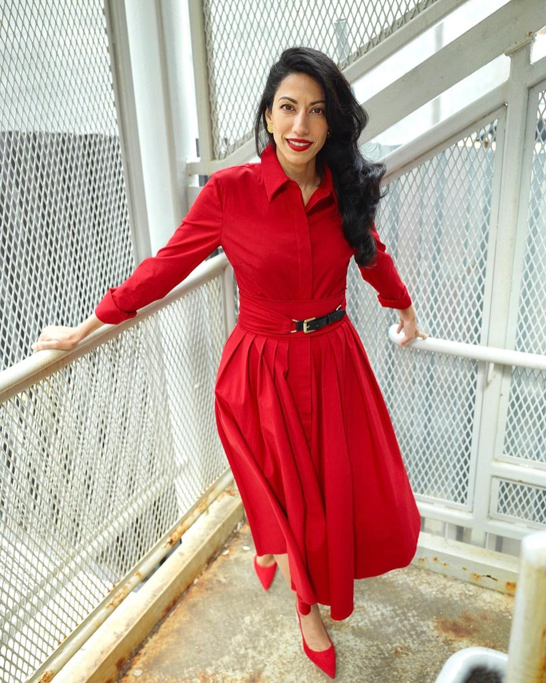 A brunette adult woman in a red dress poses on a stairway with a metal grate, showcasing a modern, vibrant fashion style.