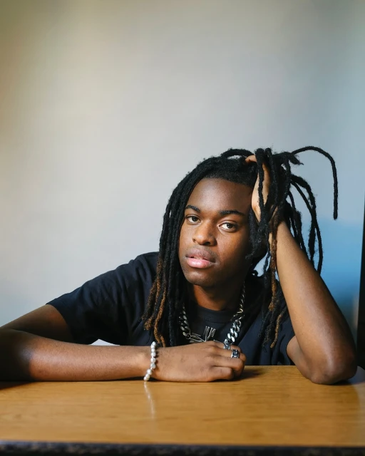 Portrait of an African-American man with dreadlocks sitting at a wooden table, wearing a black t-shirt and silver jewelry indoors.