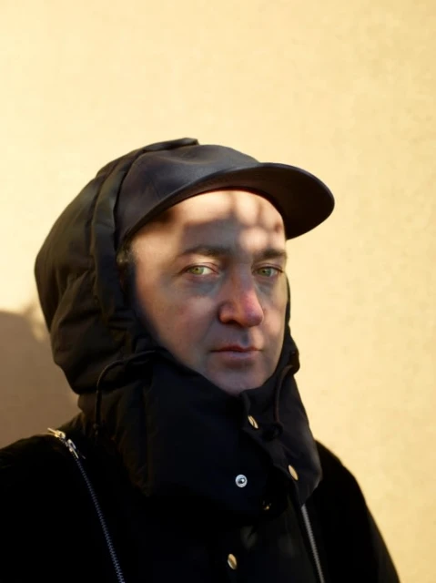 Portrait of an adult male wearing a black jacket and hat with a visor. The background is a light yellow wall with shadows.