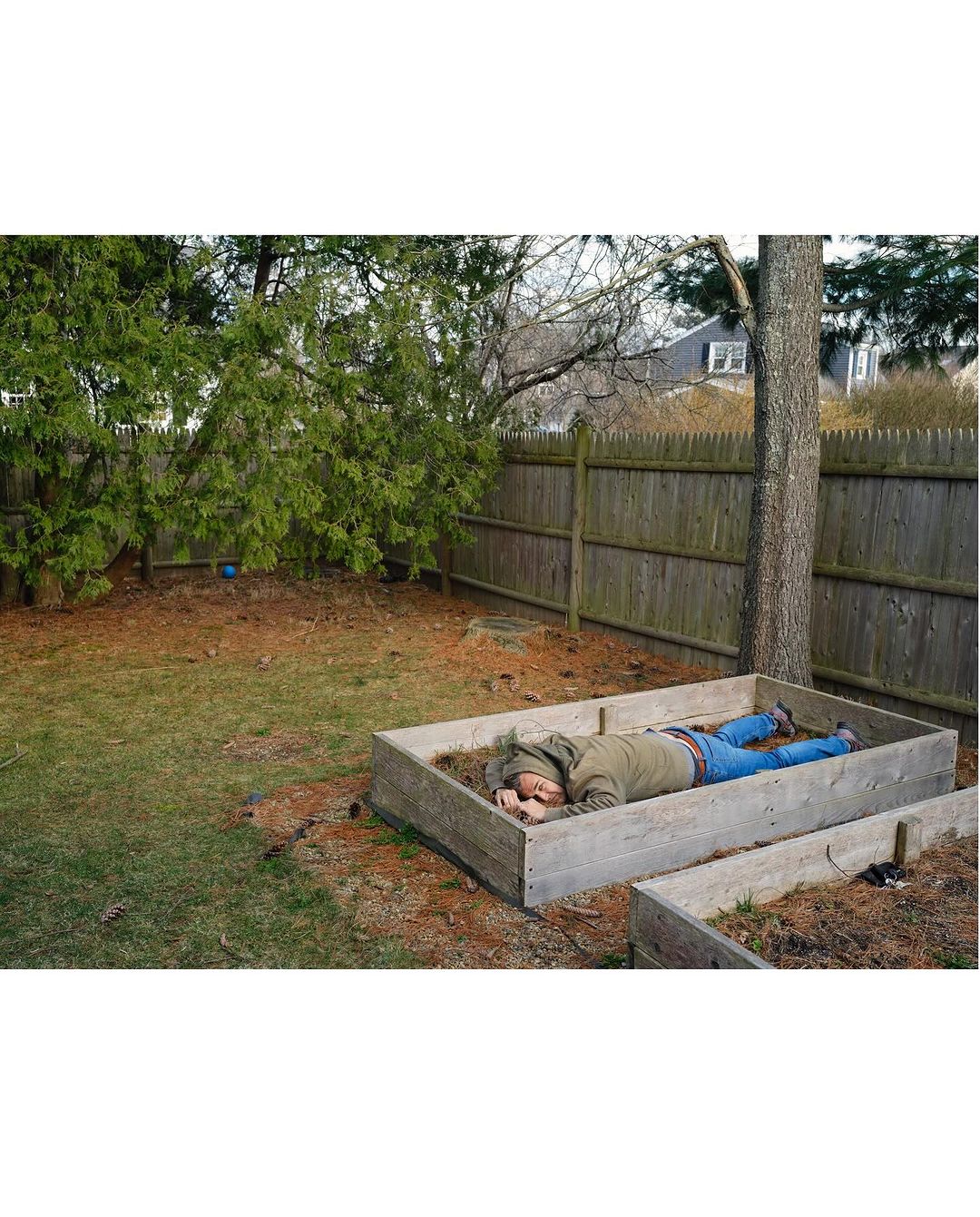 Adult male in a garden bed, wearing a hooded jacket and jeans, resting outdoors with a wooden fence and trees in the background.