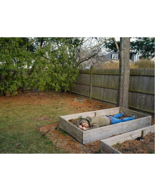 Adult male in a garden bed, wearing a hooded jacket and jeans, resting outdoors with a wooden fence and trees in the background.