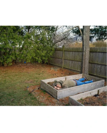 Adult male in a garden bed, wearing a hooded jacket and jeans, resting outdoors with a wooden fence and trees in the background.