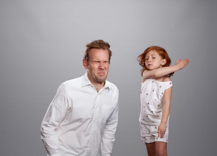 A man with a disgusted face stands next to a young girl in pajamas, who is holding her arms in a defensive pose against a gray background.