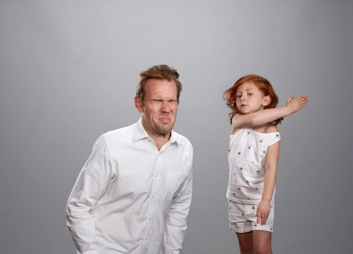 A man with a disgusted face stands next to a young girl in pajamas, who is holding her arms in a defensive pose against a gray background.