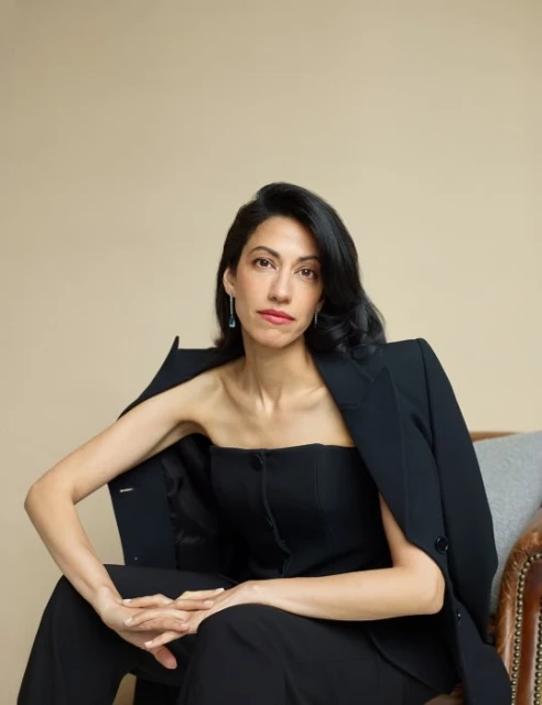 Elegant woman in black blazer and pants poses in a studio. Shoulder-length black hair, light skin, sitting indoors on a leather chair.