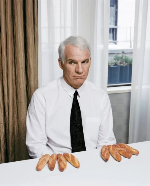 An adult male wearing a white shirt and tie sits at a table with loaves of bread on it, indoor setting with curtains and a window.