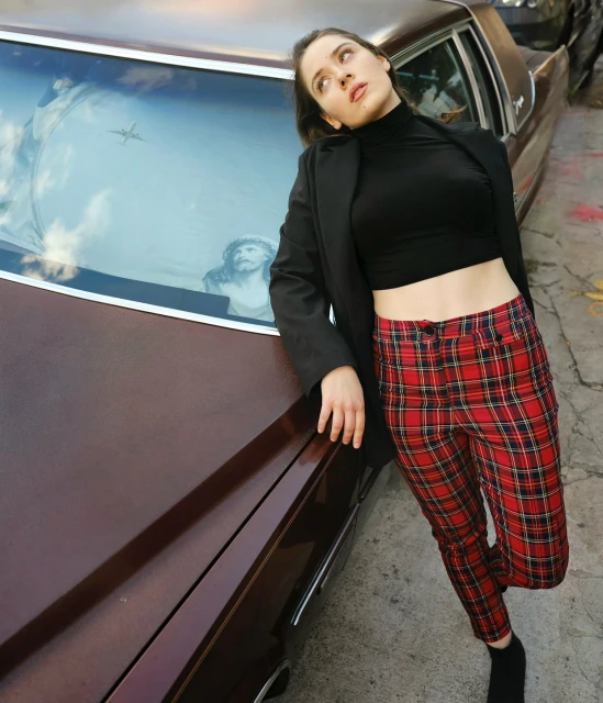 A woman poses against an old car wearing red plaid pants, black top, and blazer in an urban environment with natural lighting.
