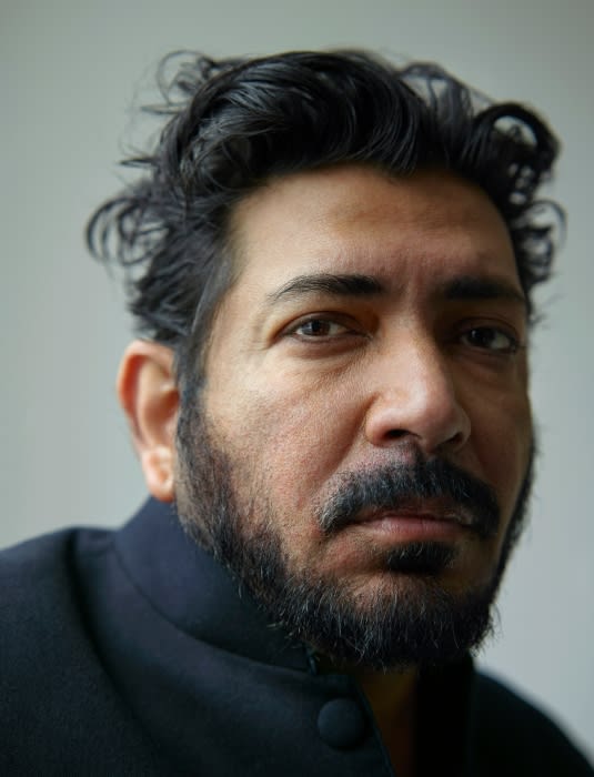 Close-up portrait of a man with dark hair, a beard, and wearing a black collared shirt against a neutral, light-colored background.