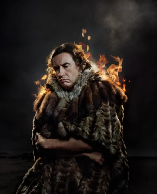 Adult male wrapped in a fur cloak, illuminated by side studio lighting and set against a dark background with visual effects of fire.