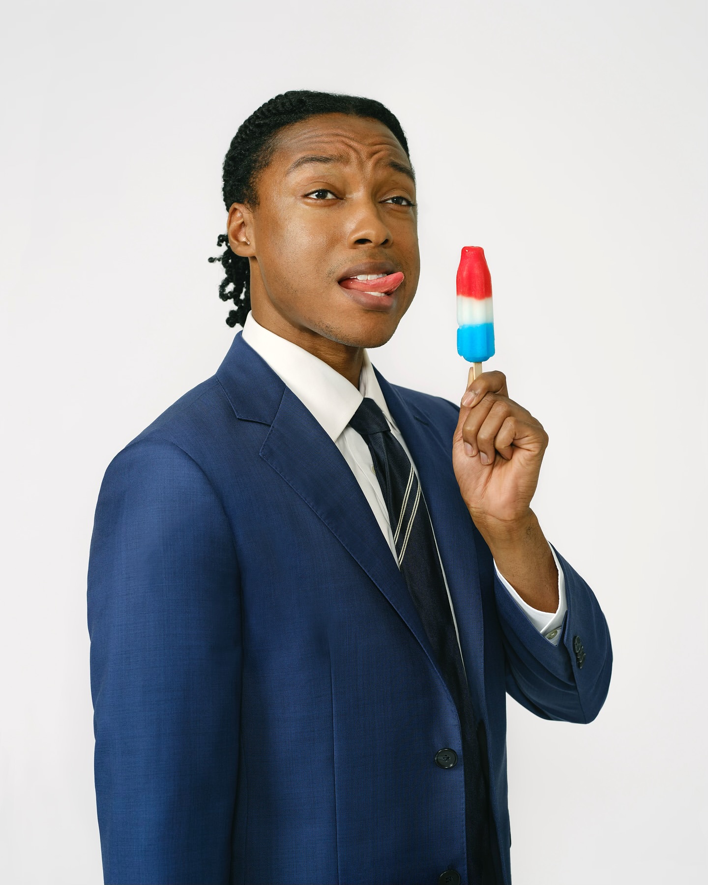 Adult male in a suit poses indoors while holding and licking a red, white, and blue ice pop with a plain white background.