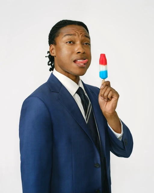 Adult male in a suit poses indoors while holding and licking a red, white, and blue ice pop with a plain white background.