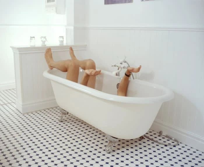 Person relaxing in a clawfoot bathtub filled with water in a bright white bathroom with patterned tile flooring.