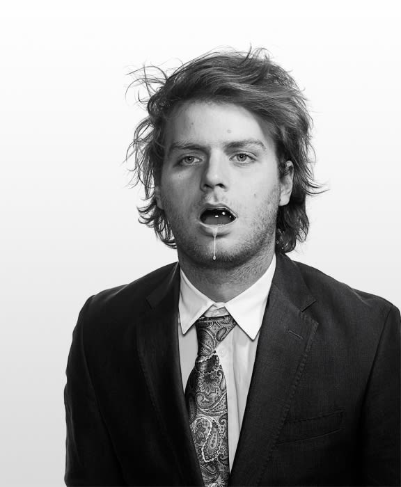 Monochrome portrait of a man in a suit with messy hair, open mouth and small white objects in mouth, drool visible, against white background.