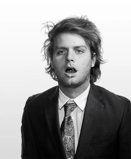 Monochrome portrait of a man in a suit with messy hair, open mouth and small white objects in mouth, drool visible, against white background.