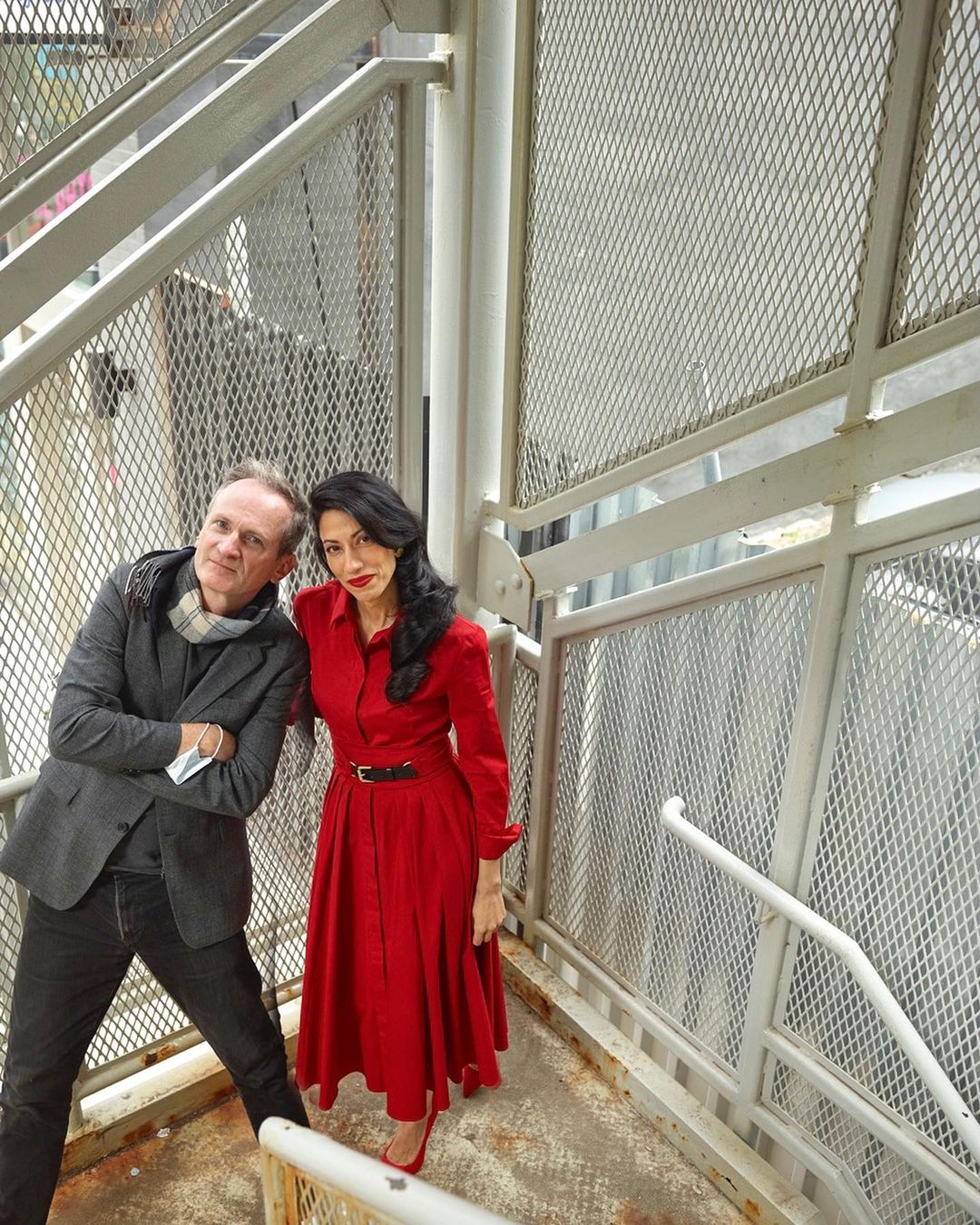 Portrait of a stylish couple posing on a metal staircase. The woman wears a red dress and heels, while the man is in a blazer and jeans.