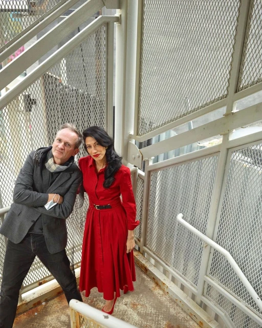 Portrait of a stylish couple posing on a metal staircase. The woman wears a red dress and heels, while the man is in a blazer and jeans.