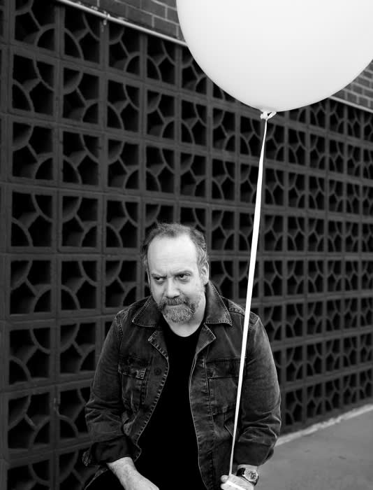 Black and white portrait of a bearded man in a denim jacket holding a white balloon against a geometric brick wall backdrop.