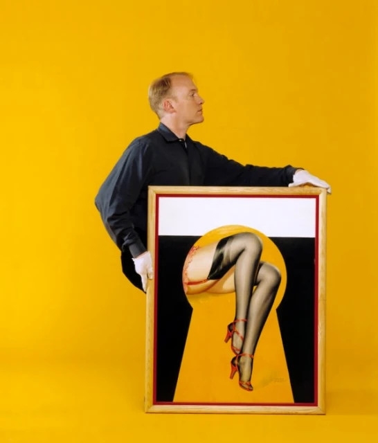 An adult man holds a framed pin-up style illustration of a woman's legs wearing red high heels in front of a bright yellow background.