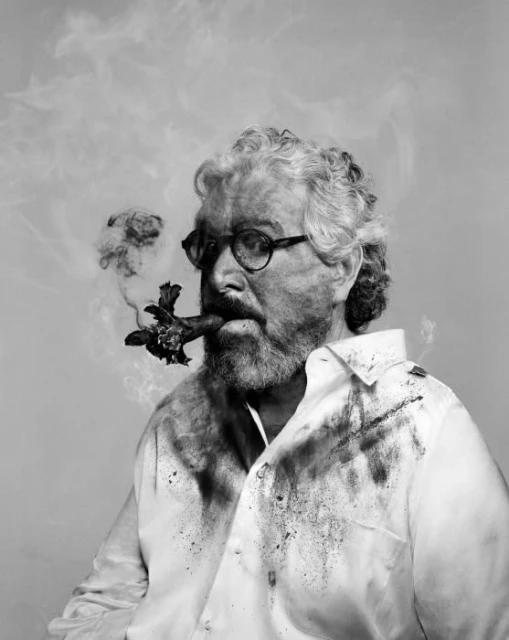 Monochrome portrait of a bearded man with eyeglasses and a white shirt smoking a cigar with dark ash smudges on his shirt.