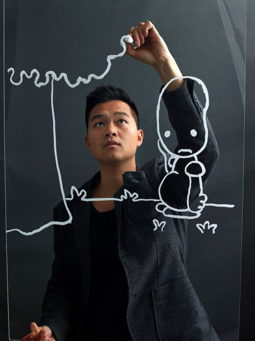 Young Asian artist in a jacket draws a cartoon figure on clear glass with a white marker against a grey background, eye-level shot.