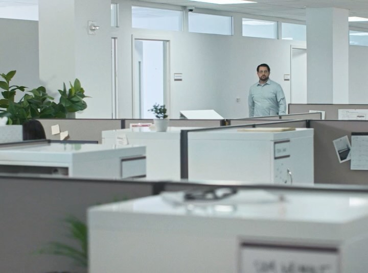 Man stands in bright office, surrounded by white cubicles with green potted plant, natural lighting and neutral color palette.