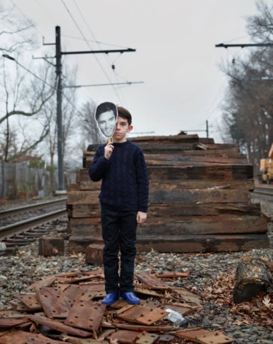 Young boy stands on railroad tracks with a male celebrity face cutout against a grey sky, wearing casual clothing and blue shoes.