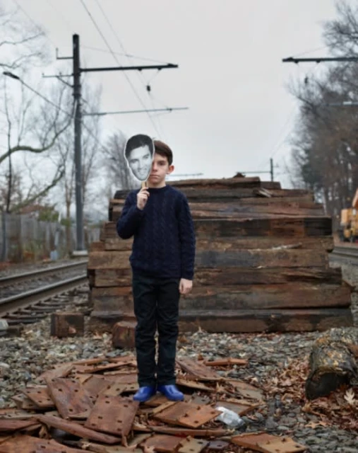 Young boy stands on railroad tracks with a male celebrity face cutout against a grey sky, wearing casual clothing and blue shoes.