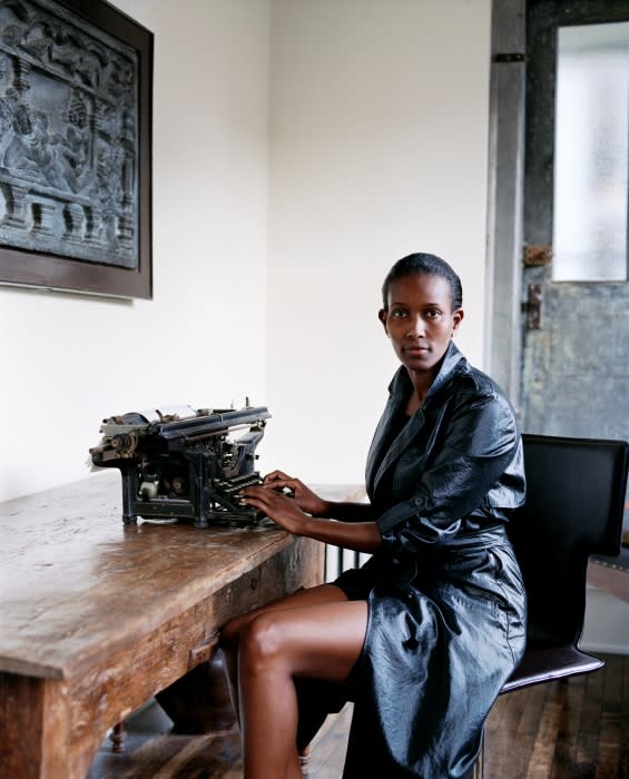 Stylish woman in shiny black robe sits at a wooden desk at a vintage typewriter in an indoor setting with a decorative background.