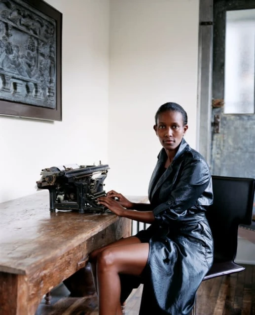 Stylish woman in shiny black robe sits at a wooden desk at a vintage typewriter in an indoor setting with a decorative background.