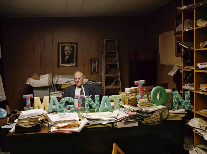 Elderly man in suit sits behind a cluttered desk with large 'Imagination' letters, surrounded by paperwork, in an office space.