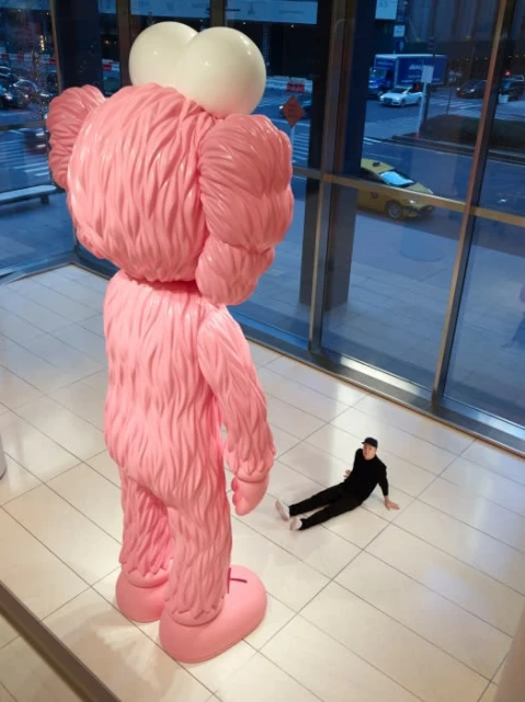 Large, pink KAWS sculpture in an indoor setting, with a casually posed person in black alongside the oversized, eye-catching artwork.
