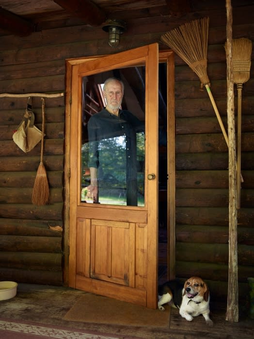 An elderly man stands in an open wooden door of a log cabin, peering out at the camera. A beagle dog rests on the porch.
