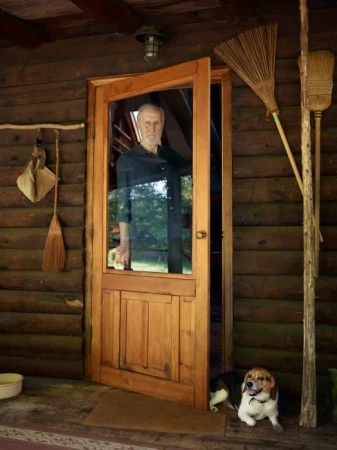 An elderly man stands in an open wooden door of a log cabin, peering out at the camera. A beagle dog rests on the porch.