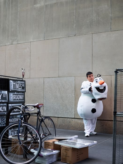 Man in Olaf costume holds head in urban setting with bicycles and street signs. Building facade backdrop with muted color palette.