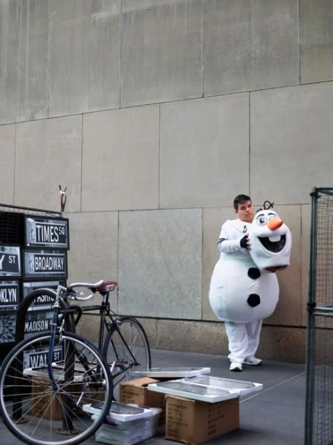 Man in Olaf costume holds head in urban setting with bicycles and street signs. Building facade backdrop with muted color palette.