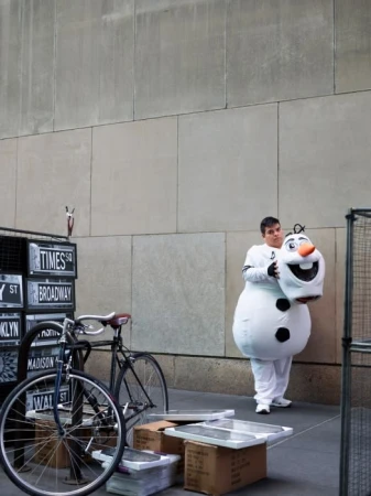 Man in Olaf costume holds head in urban setting with bicycles and street signs. Building facade backdrop with muted color palette.