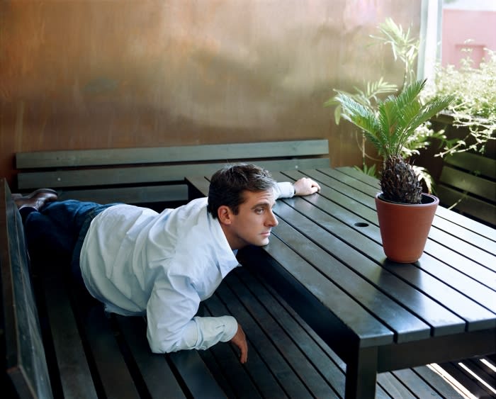 Man in a dress shirt and jeans lying on an outdoor bench next to a table with a potted palm plant, looking off to the side.
