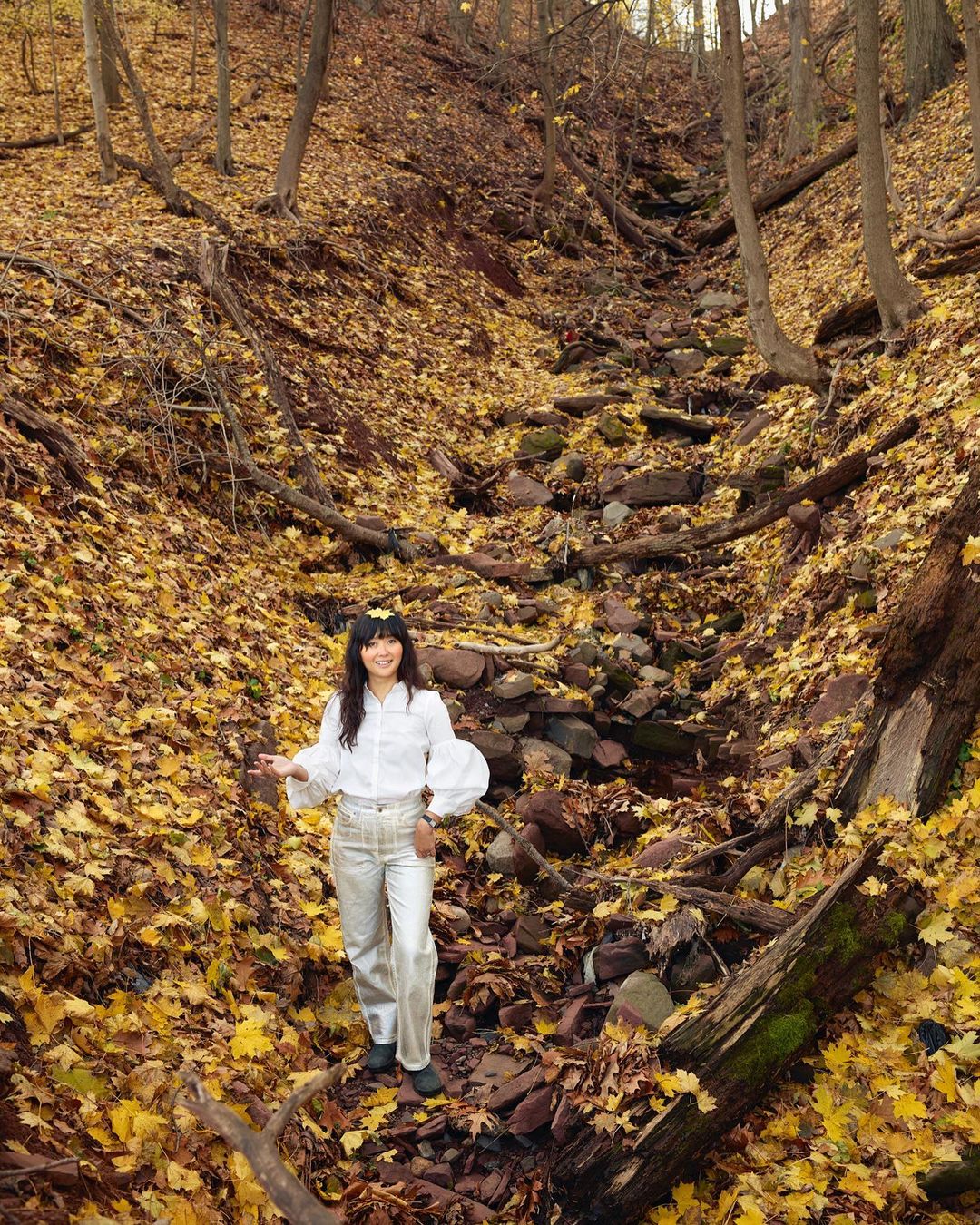 Woman stands amidst a forest floor covered with fallen yellow leaves in autumn, smiling beneath a canopy of bare trees.