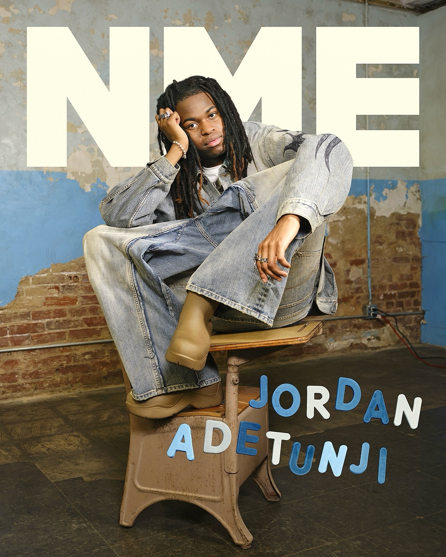 Young man with dreadlocks wearing a denim outfit sits on a stool in a room with brick walls and a large NME logo in this studio portrait.
