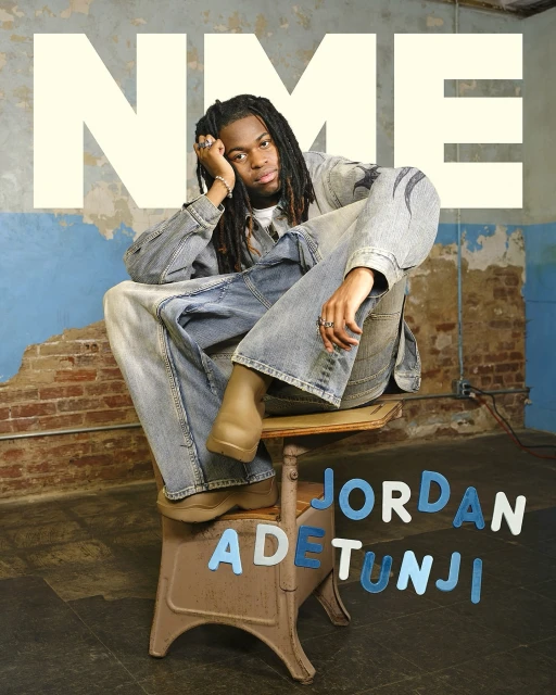 Young man with dreadlocks wearing a denim outfit sits on a stool in a room with brick walls and a large NME logo in this studio portrait.