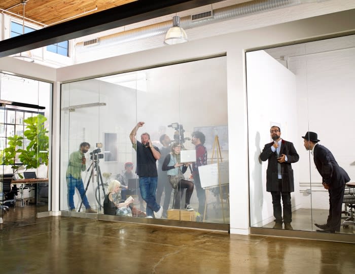 Behind glass windows, a video and photo shoot in progress featuring a diverse group of adults, with two businessmen observing.