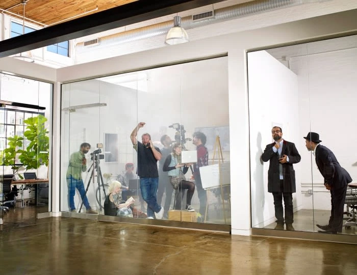 Behind glass windows, a video and photo shoot in progress featuring a diverse group of adults, with two businessmen observing.